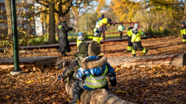 Två barn sitter på en liggande trädstock i en park och barn i bakgrunden leker på lekställningar.